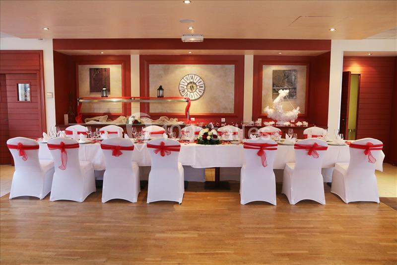 Salle de réception avec une longue table décorée de nappes blanches et de nœuds rouges sur les chaises.