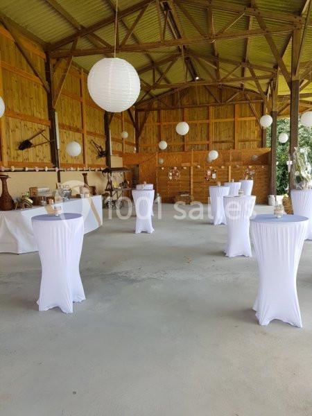 Salle de réception rustique avec tables hautes drapées de blanc, lanternes suspendues et décorations en bois.