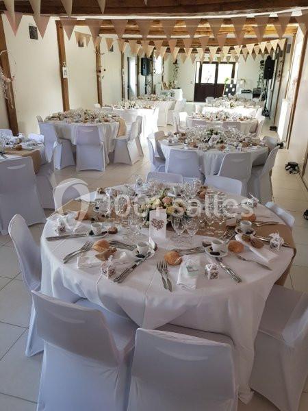 Salle de réception décorée pour un mariage, avec tables rondes dressées, nappes blanches et guirlandes suspendues.
