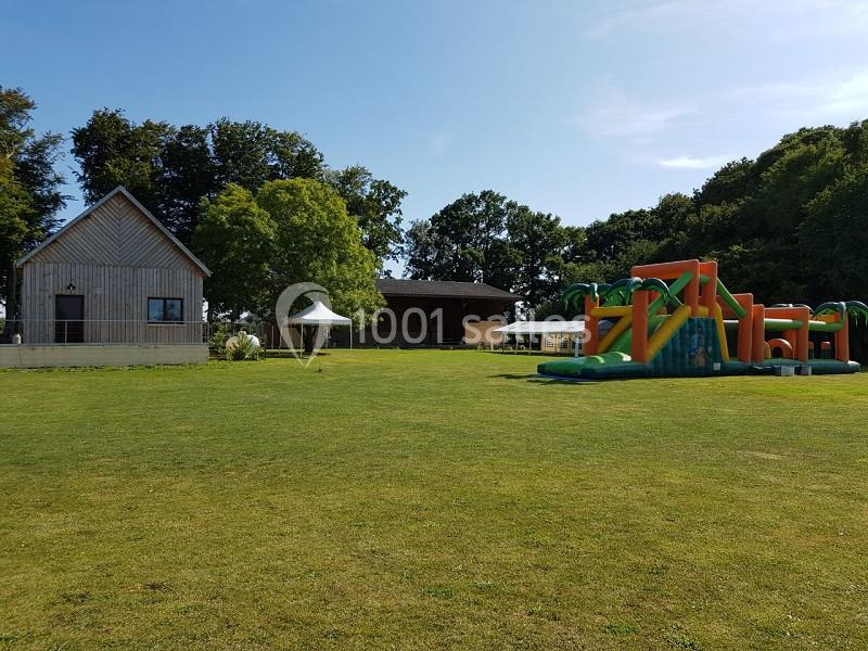 Grande pelouse avec structure gonflable colorée, bâtiment en bois et arbres en arrière-plan sous un ciel dégagé.
