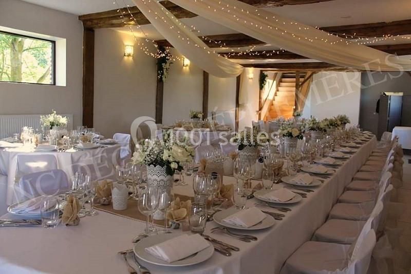 Salle de réception décorée avec des tables alignées, nappes blanches, vaisselle élégante et guirlandes lumineuses suspendues.