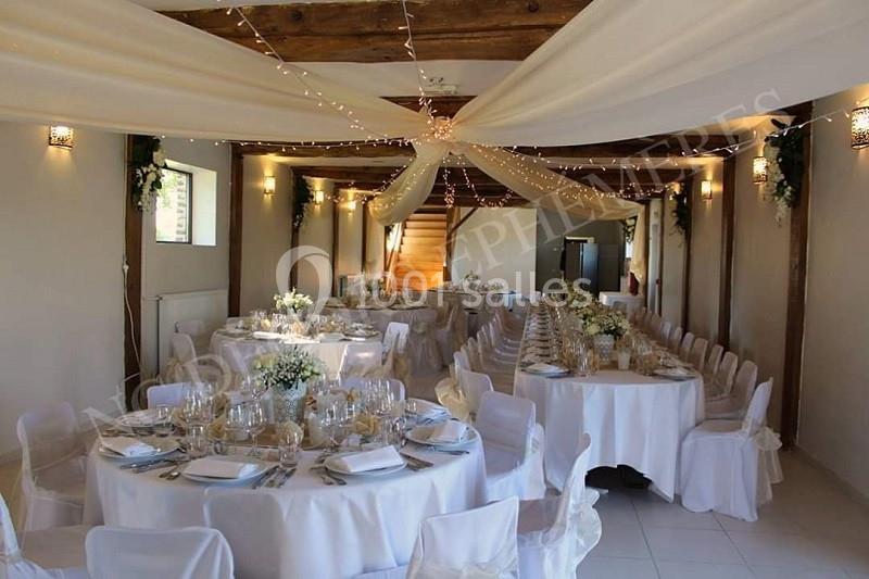 Salle décorée pour un mariage avec tables rondes dressées, nappes blanches, fleurs et guirlandes lumineuses suspendues.