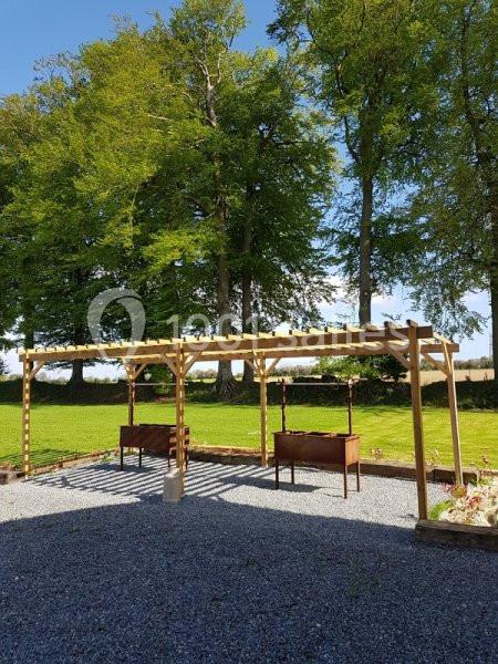 Pergola en bois avec deux ruches en dessous, située sur un sol gravillonné devant un espace vert et des arbres.