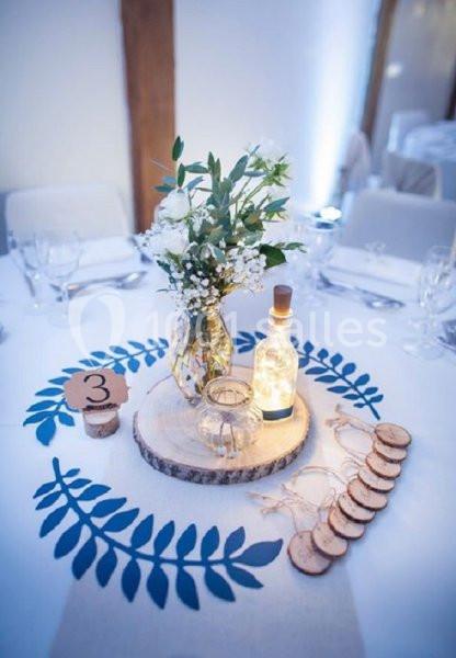 Centre de table avec vase fleuri, bouteille lumineuse, rondins de bois et numéro de table sur une nappe blanche décorée.