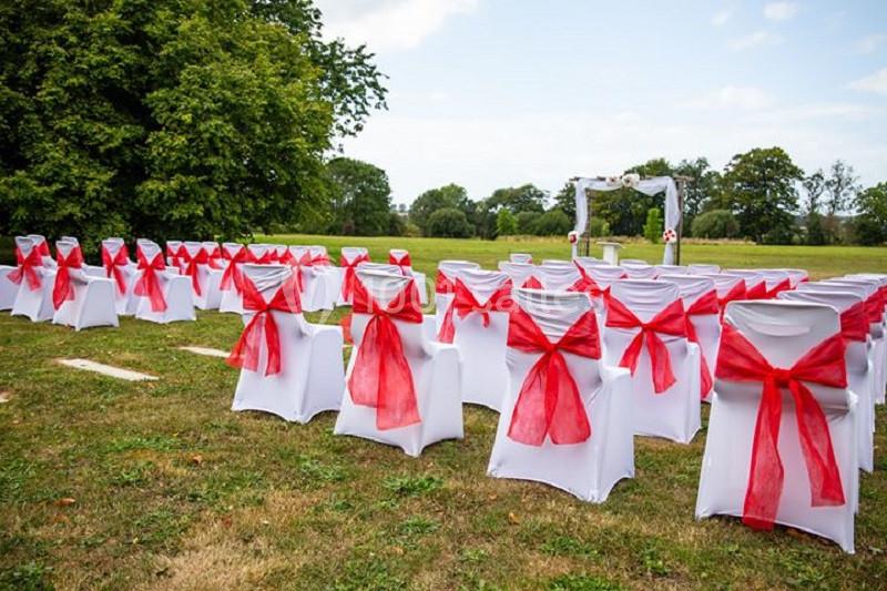Chaises blanches décorées de nœuds rouges alignées en extérieur, avec une arche de mariage en arrière-plan.