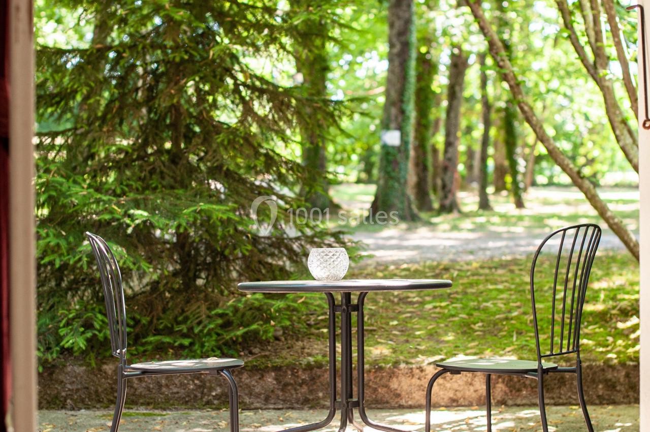 Table et deux chaises en métal noir sur une terrasse ombragée, entourées d'arbres dans un cadre naturel verdoyant.