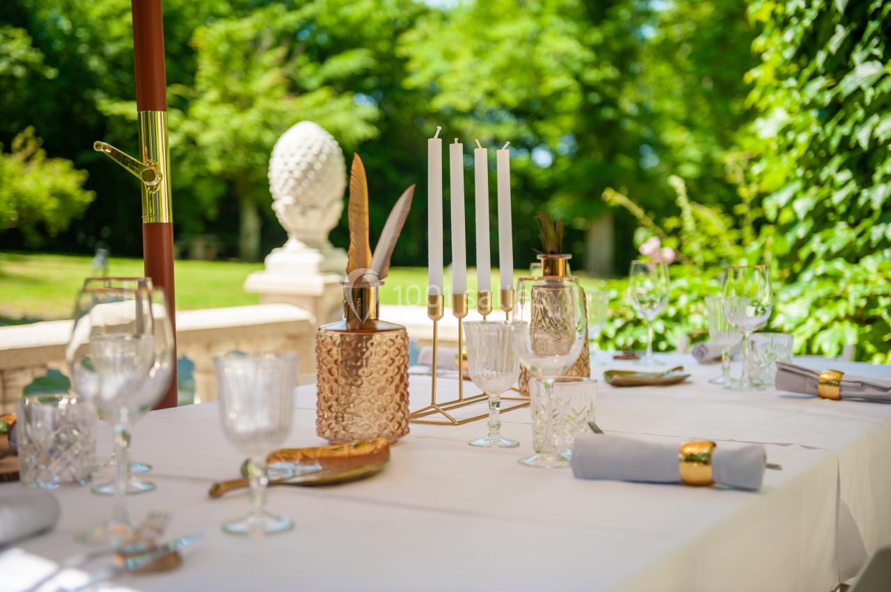 Table élégamment dressée en extérieur avec bougeoirs, verres en cristal et vue sur un jardin verdoyant.