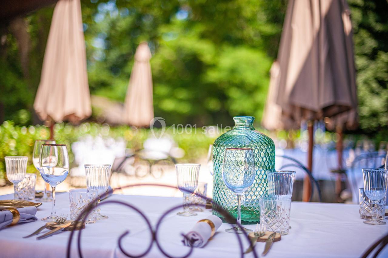 Table dressée en extérieur avec une carafe en verre vert, des verres et des serviettes, sous des parasols beige.