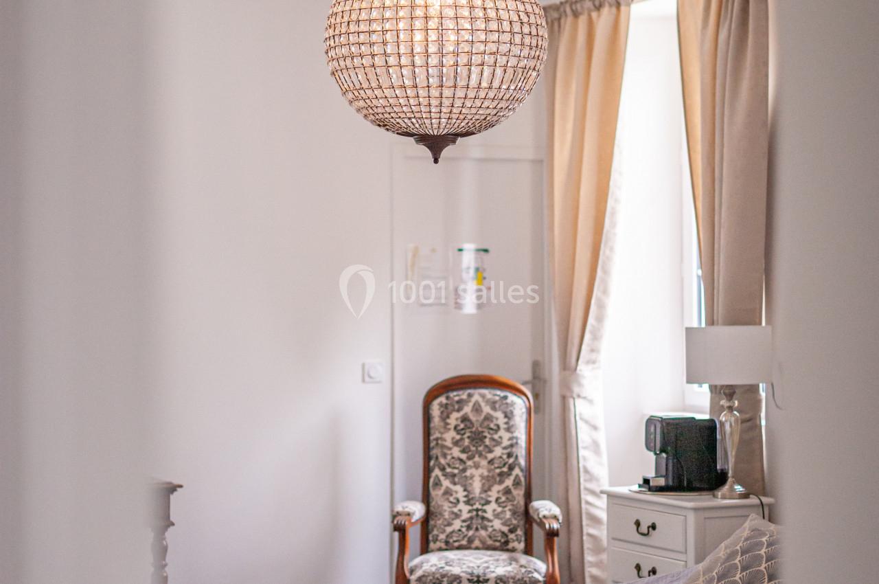 Chambre lumineuse avec un lit, un fauteuil, une commode et un lustre en perles, décorée dans des tons clairs et neutres.