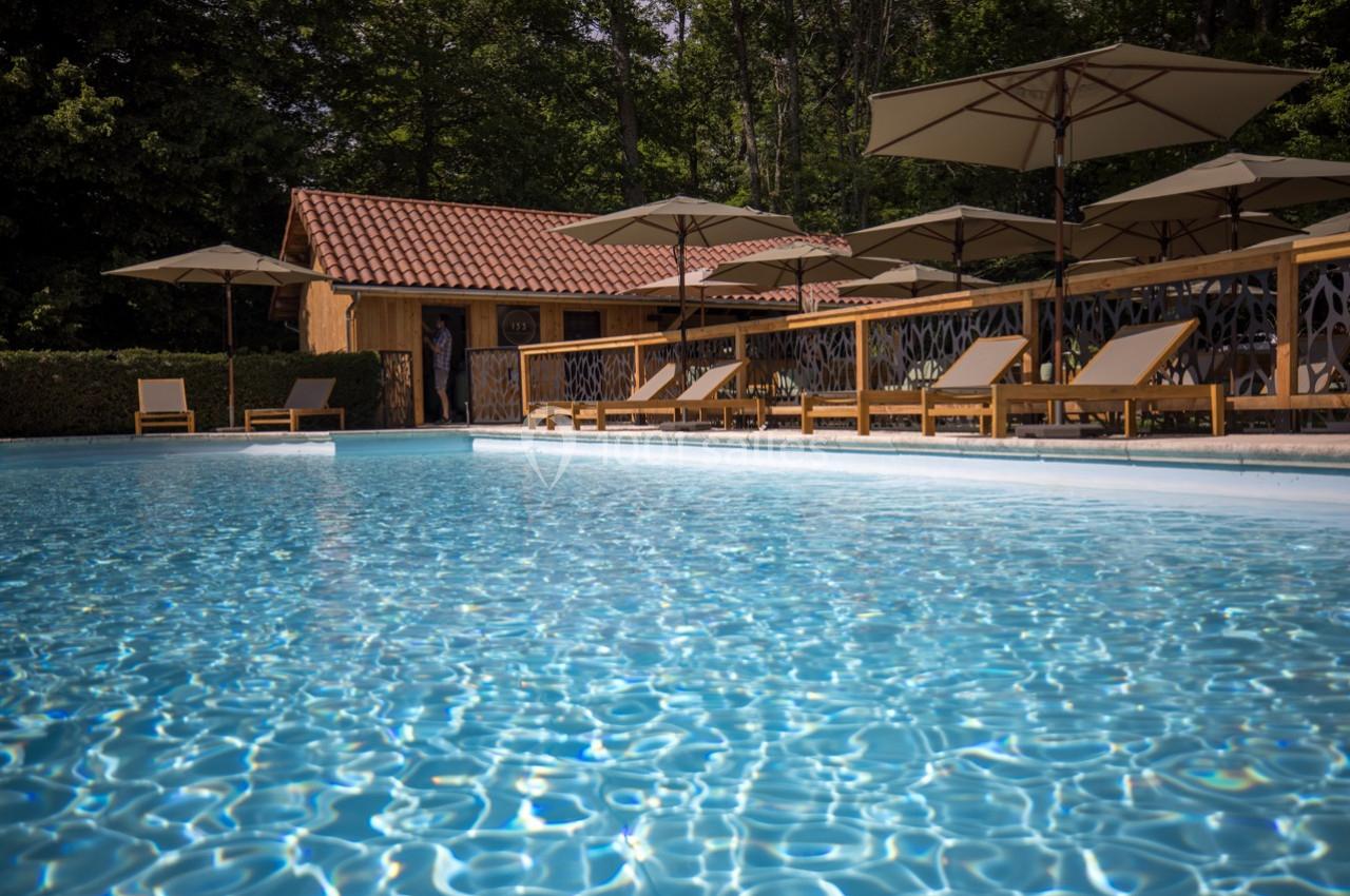 Piscine extérieure entourée de chaises longues, parasols et cabanon en bois, bordée par des arbres.