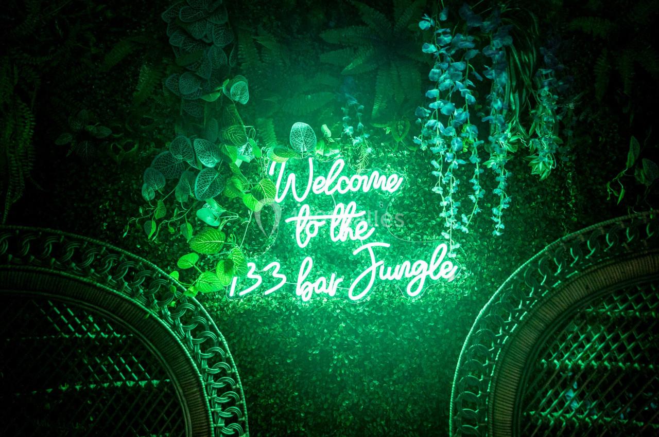 Néon vert indiquant ’Welcome to the 133 bar Jungle’ sur un mur végétal décoré de feuillages et de fleurs.