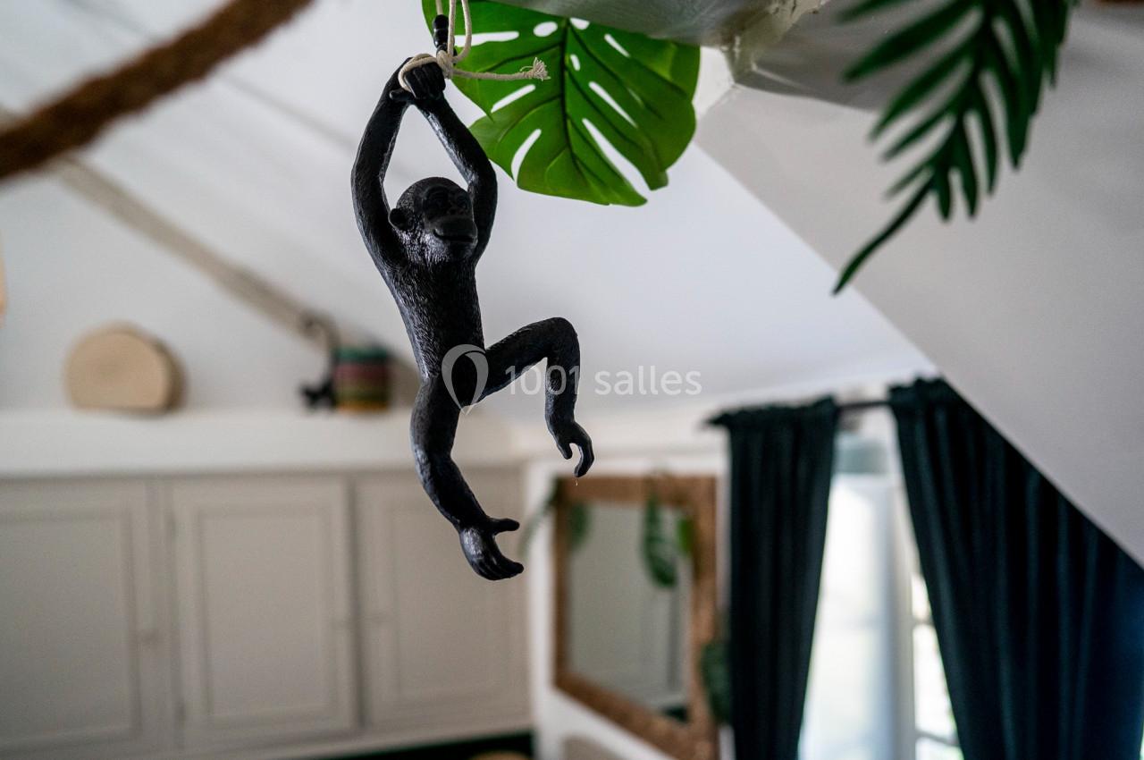 Sculpture de singe noir suspendue à une corde sous un plafond incliné, entourée de feuilles décoratives.