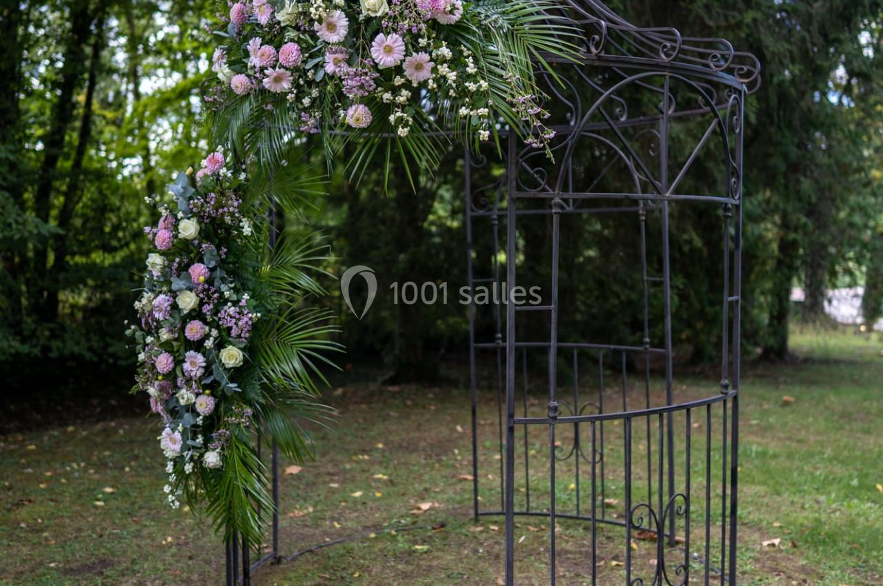 Structure métallique décorée de fleurs roses et blanches dans un jardin entouré d'arbres.