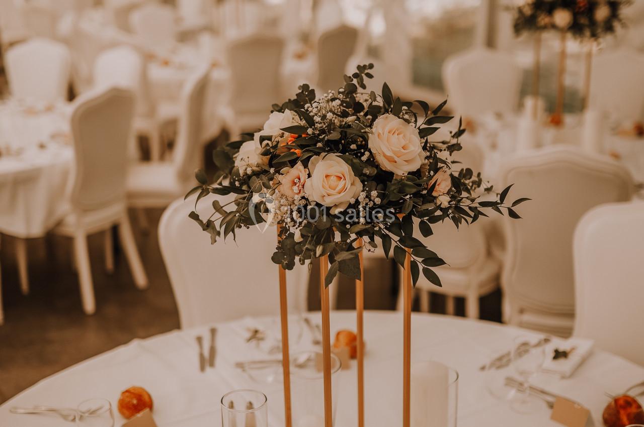Salle de réception décorée avec des tables rondes, centres de table floraux et chaises blanches sous une tente.