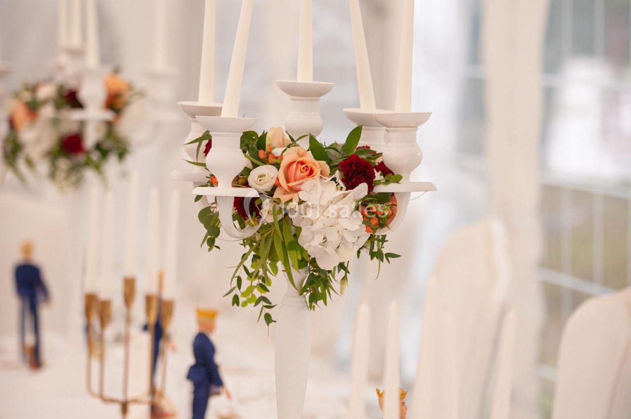 Bougeoirs blancs ornés de fleurs au centre d'une table de réception élégamment dressée sous une tente lumineuse.