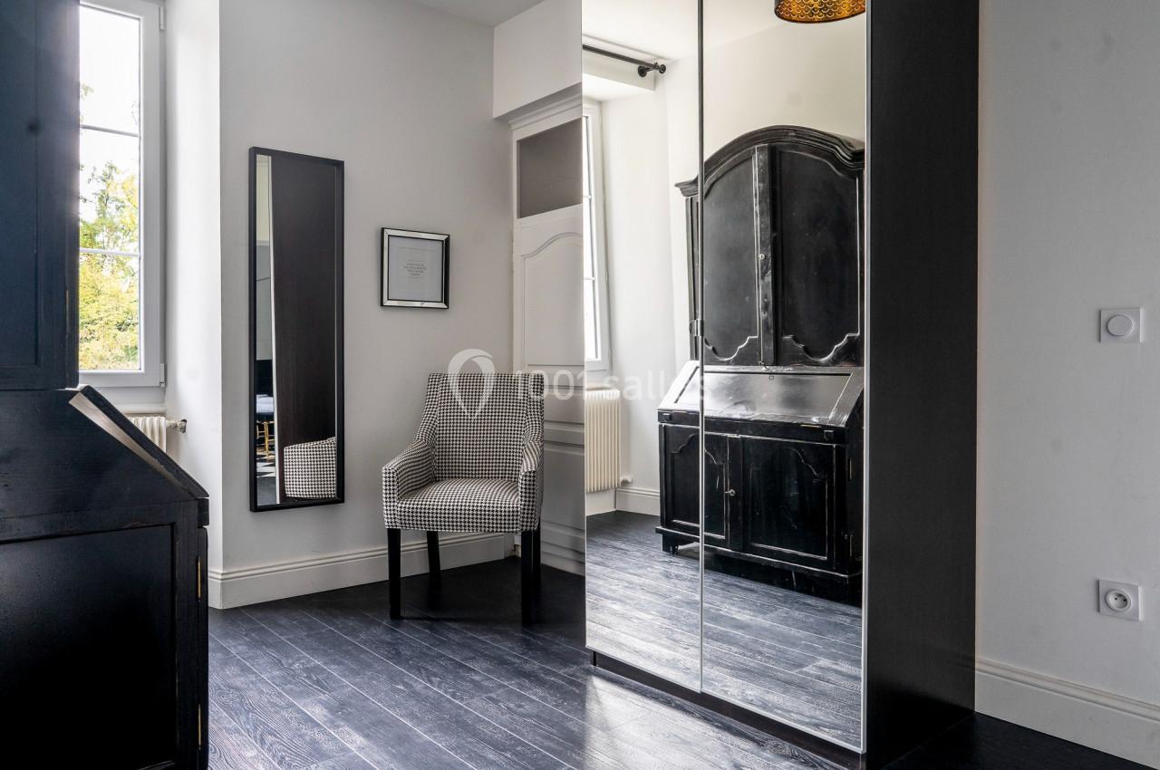 Chambre lumineuse avec parquet sombre, armoire noire, miroir, fauteuil à motif et fenêtre laissant entrer la lumière.