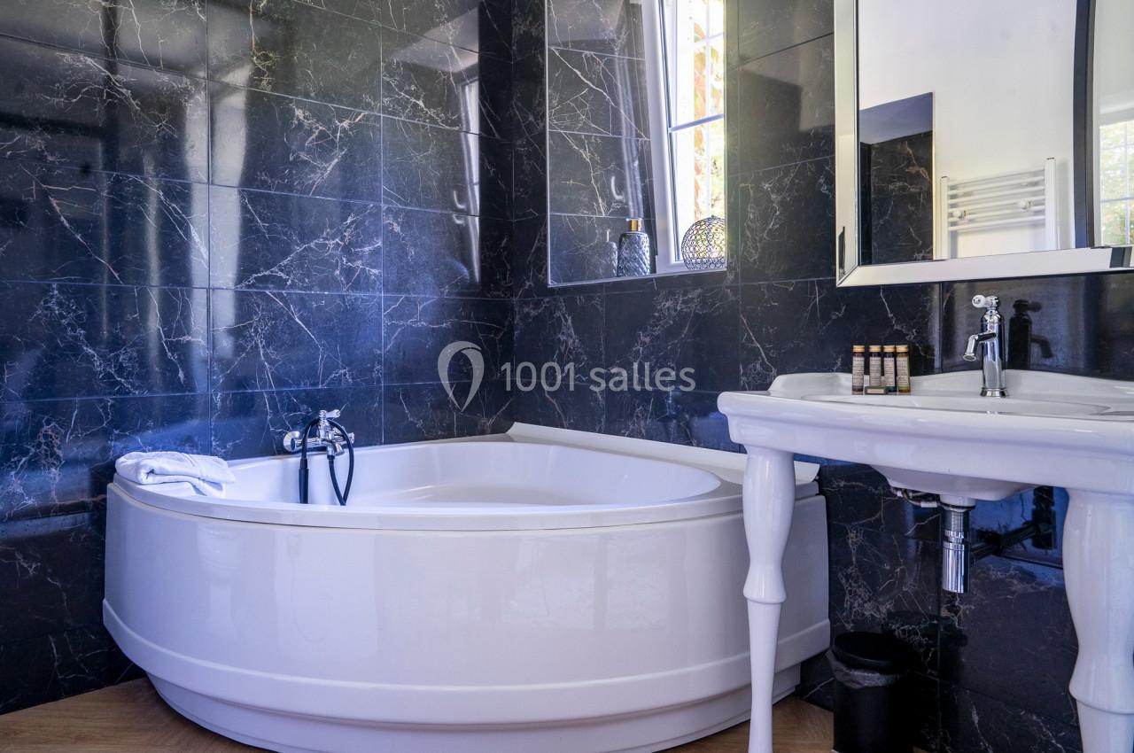 Salle de bain avec baignoire d'angle blanche, murs en marbre noir, lavabo sur pied et fenêtre laissant entrer la lumière.