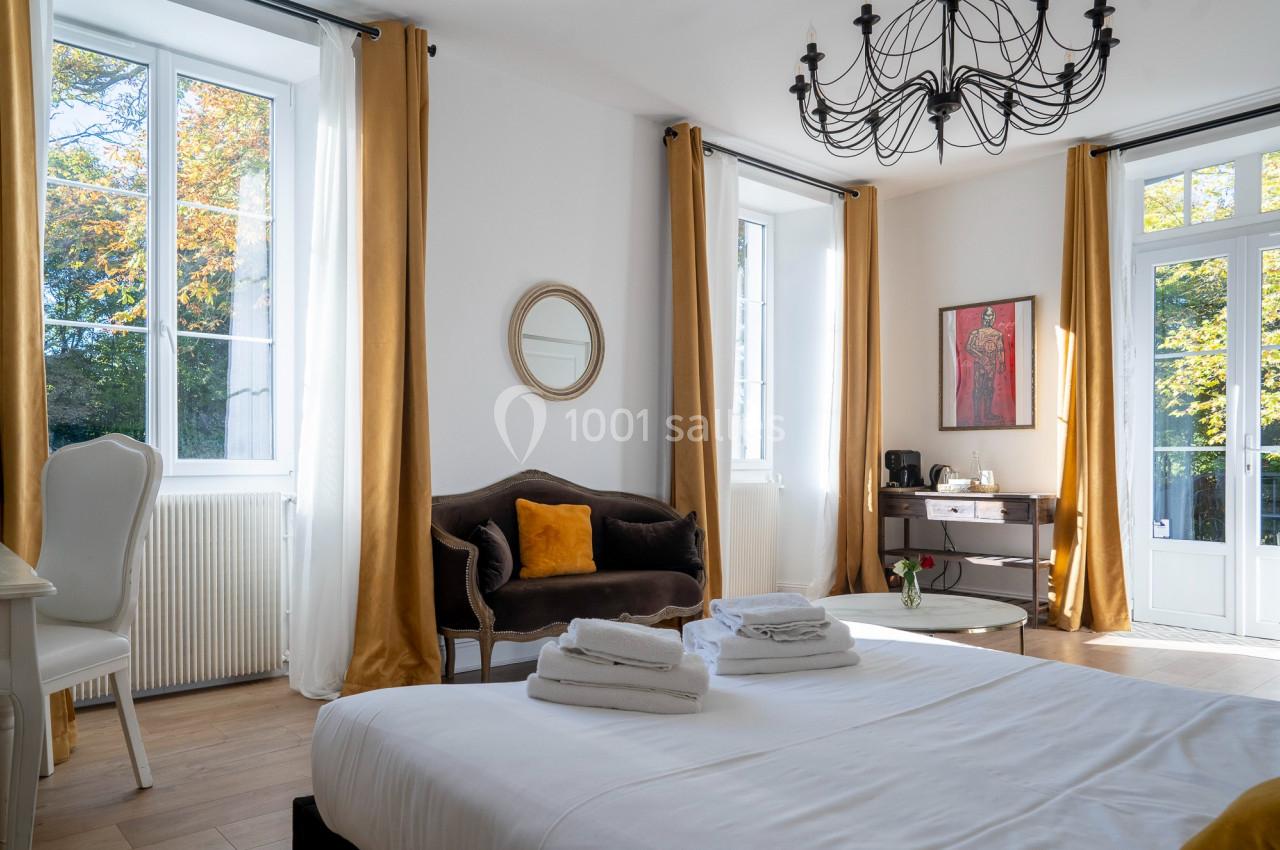 Chambre lumineuse avec lit double, fauteuil, rideaux dorés, lustre noir et vue sur un jardin à travers de grandes fenêtres.