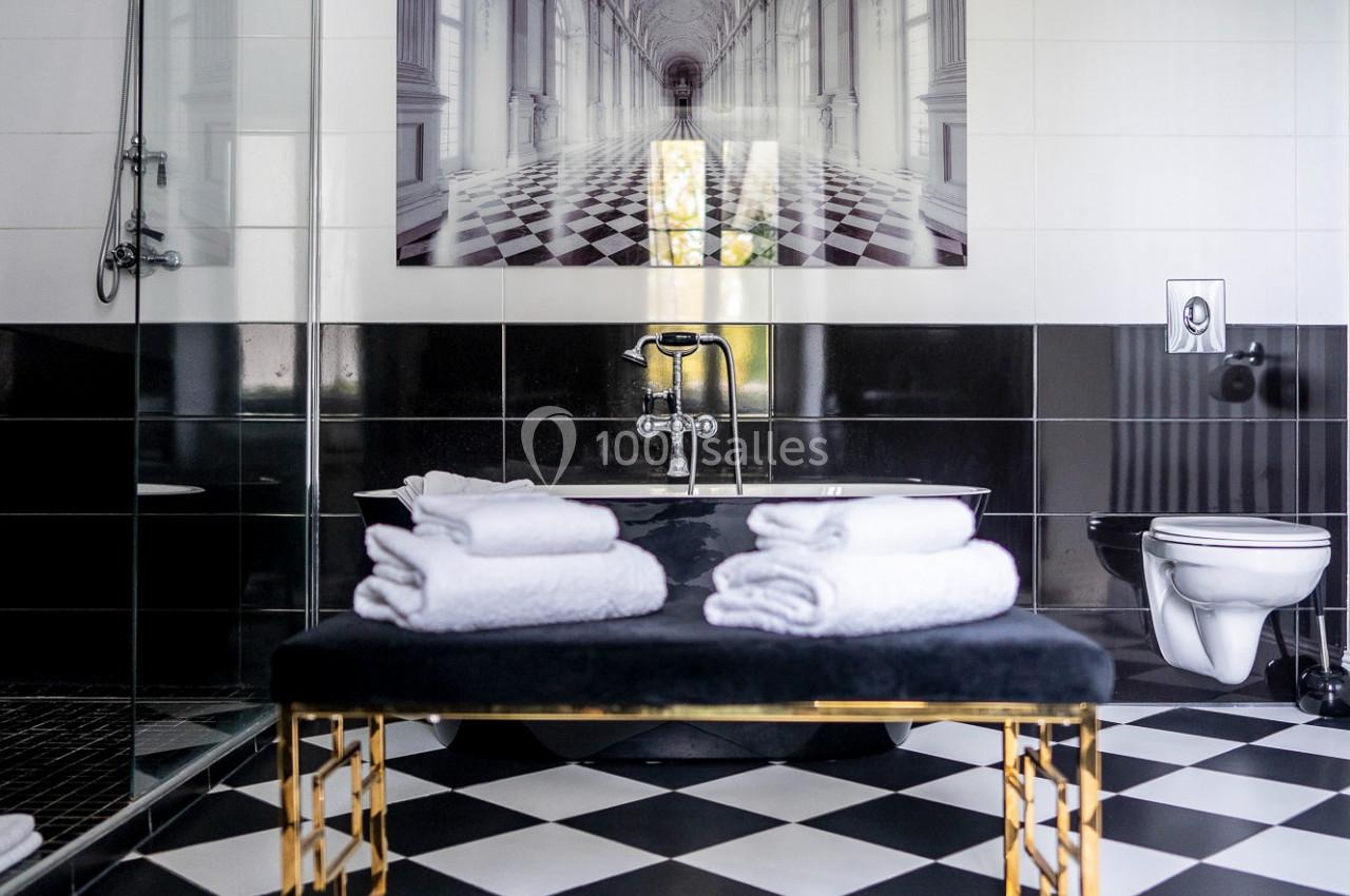 Salle de bain moderne avec carrelage noir et blanc, douche vitrée, banc doré et serviettes pliées.