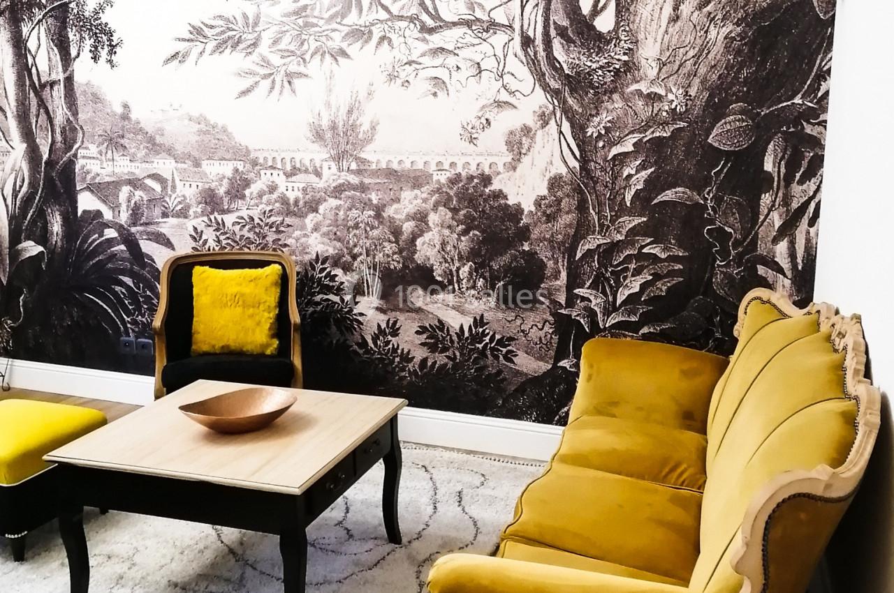 Salon avec canapé et fauteuils jaunes, table basse en bois, tapis clair et mur décoré d'une fresque végétale en noir et…