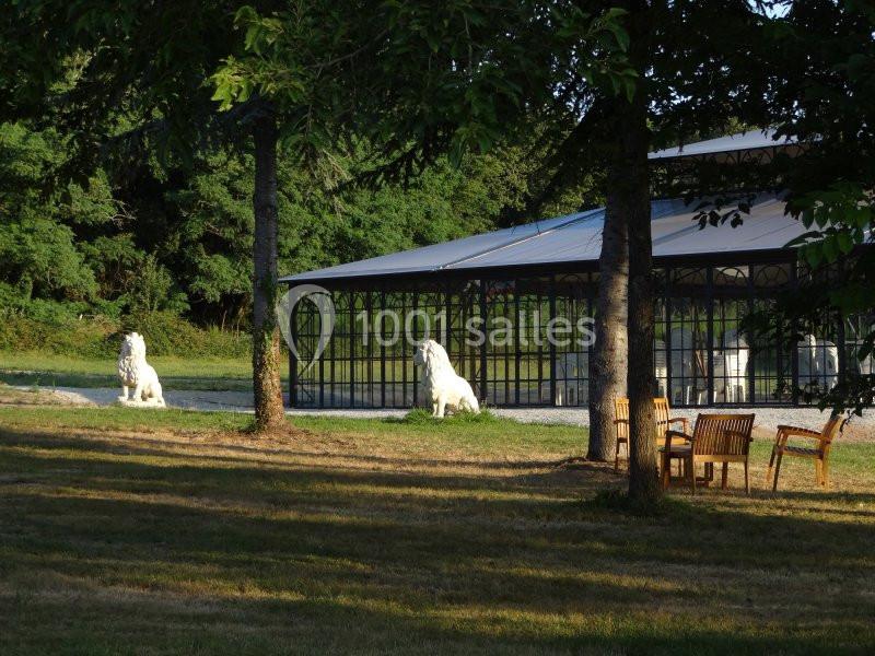 Deux statues de lions blancs devant une verrière, entourées d'arbres et de mobilier de jardin en bois.