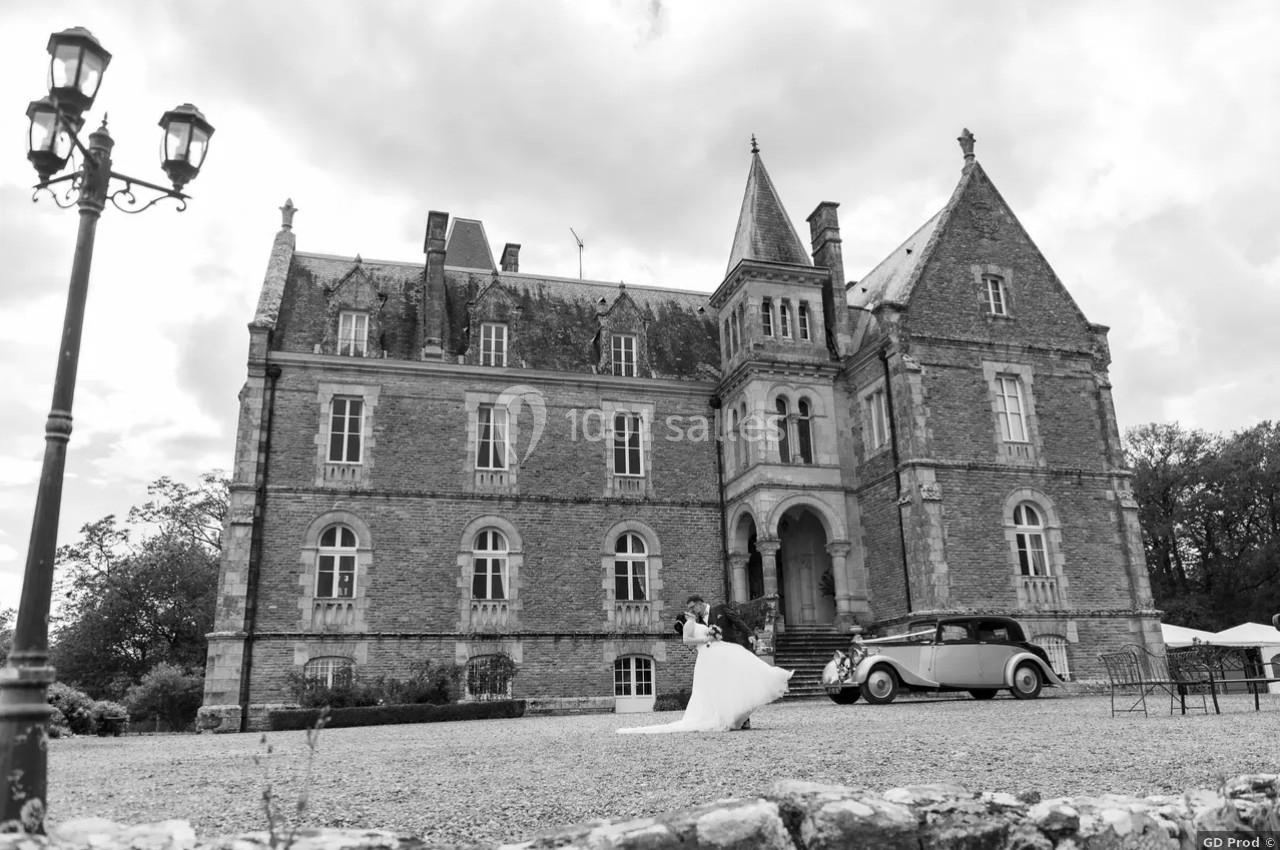 Un couple de mariés pose devant un château en pierre, avec une voiture ancienne stationnée à proximité.