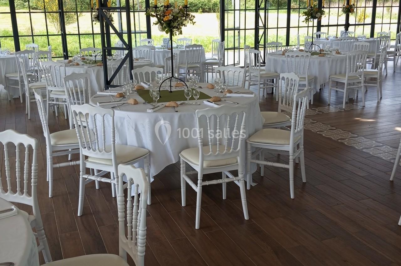 Salle de réception lumineuse avec tables rondes dressées, nappes blanches et chaises blanches, vue sur un jardin.