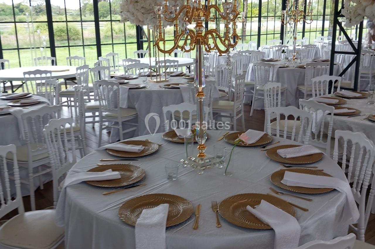 Salle de réception décorée avec des tables rondes dressées, nappes blanches, chandeliers dorés et chaises blanches.
