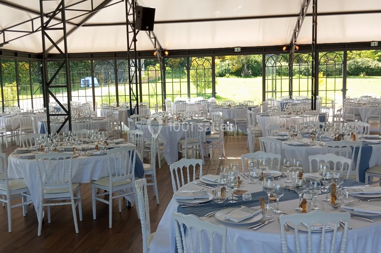 Salle de réception lumineuse avec tables rondes dressées, chaises blanches et vue sur un jardin à travers des baies vitrées.