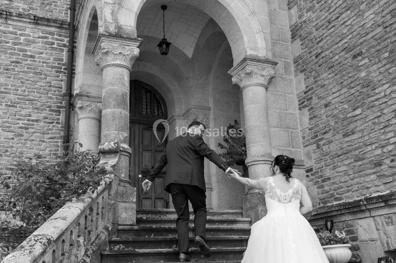 Un couple monte les marches d'un bâtiment ancien en pierre, la mariée en robe blanche tenant la main du marié.