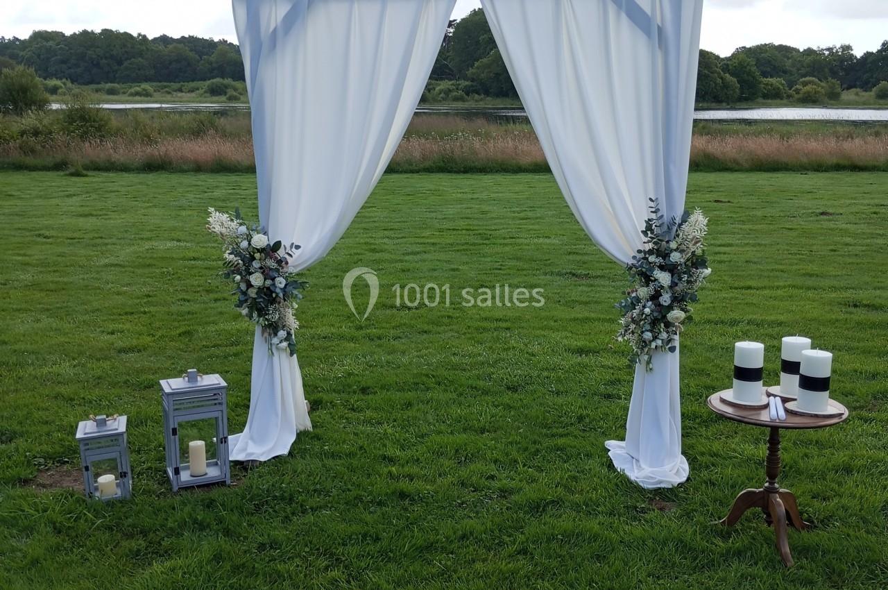 Arche de cérémonie en bois avec rideaux blancs, décorée de fleurs, sur une pelouse près d'un plan d'eau.