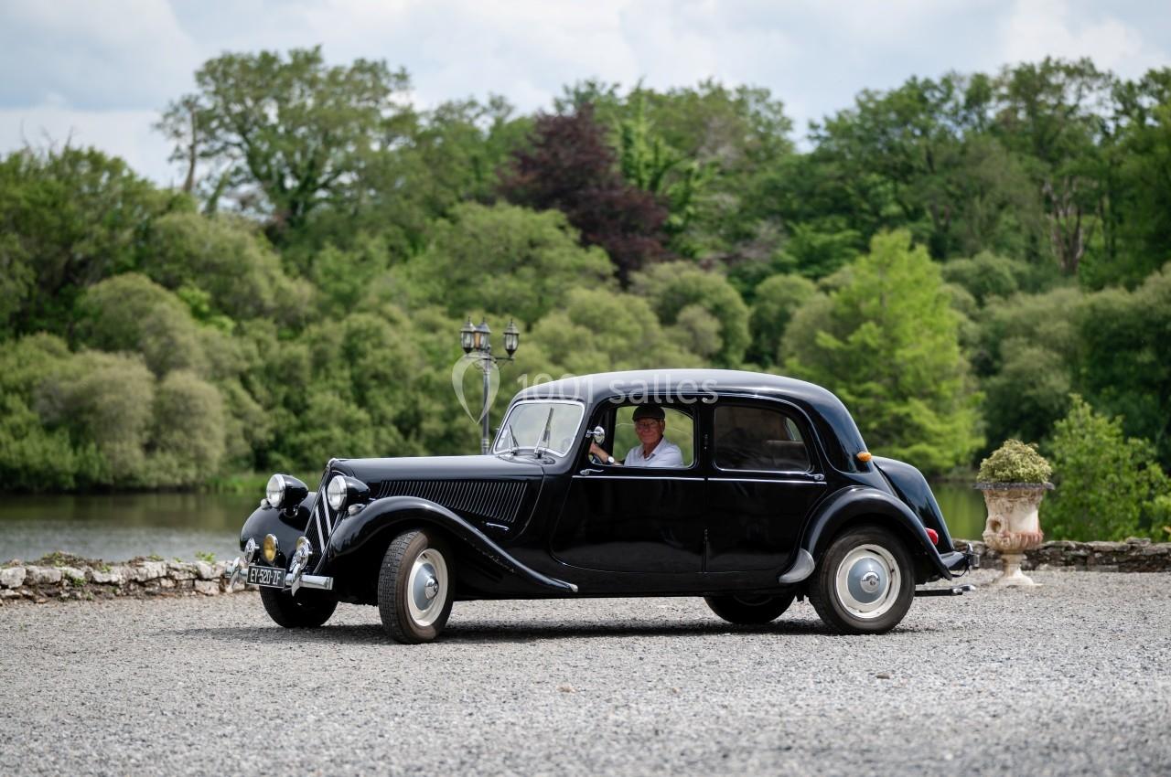 Voiture ancienne noire stationnée sur un chemin gravillonné, avec un conducteur visible, entourée de verdure.
