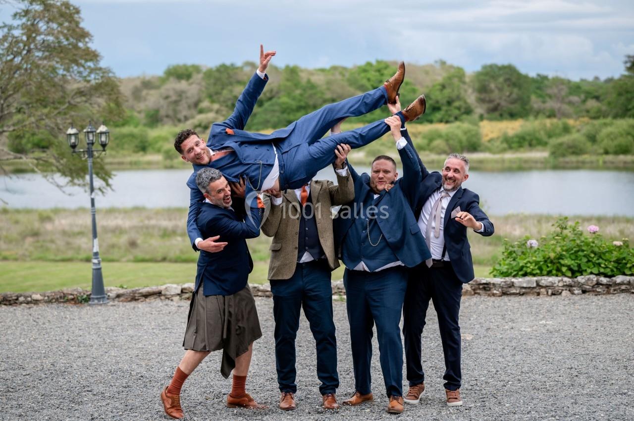 Cinq hommes en tenue formelle, souriants, soulèvent un ami en costume bleu devant un paysage naturel avec un lac.