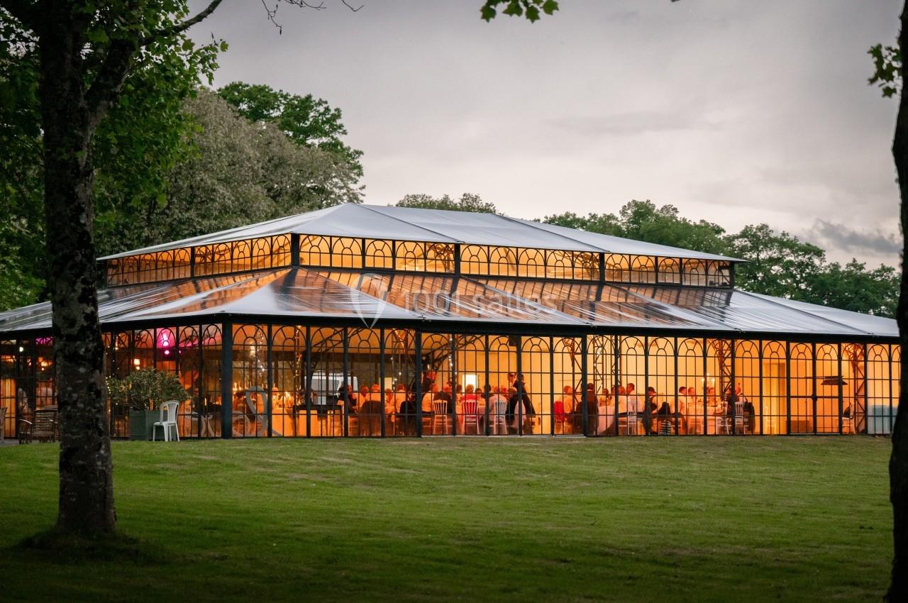 Grande verrière éclairée en soirée, entourée d'arbres et située sur une pelouse verdoyante.