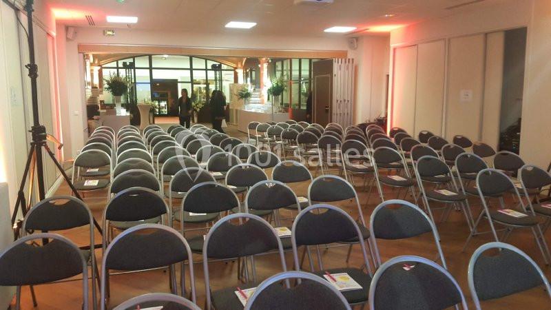 Salle de conférence avec des rangées de chaises vides, éclairage chaleureux et vue sur une entrée vitrée.