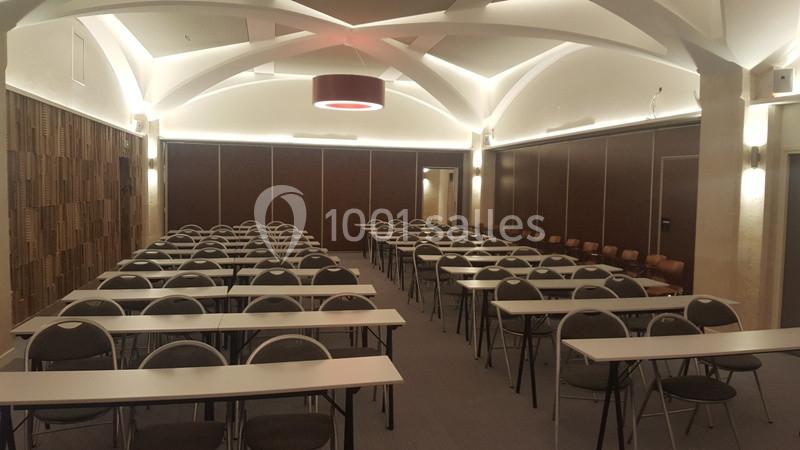Salle de conférence avec des rangées de tables et chaises alignées, éclairée par des lumières au plafond voûté.