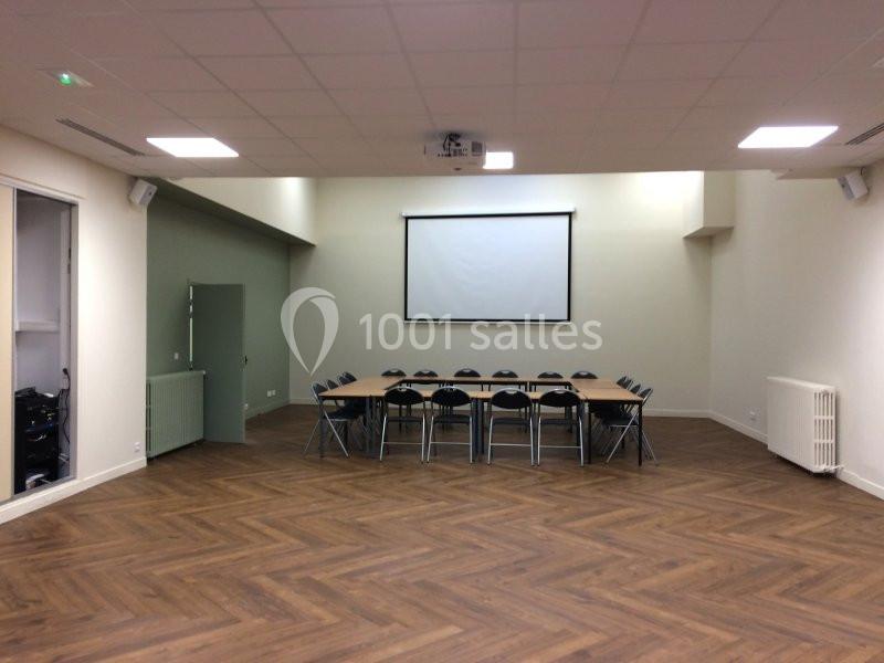 Salle de réunion vide avec table rectangulaire, chaises pliantes, parquet en bois et écran de projection au mur.