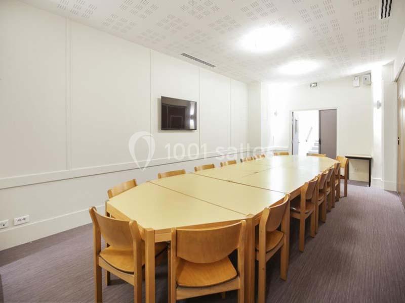 Salle de réunion lumineuse avec une grande table en bois entourée de chaises, un écran mural et un sol en moquette.