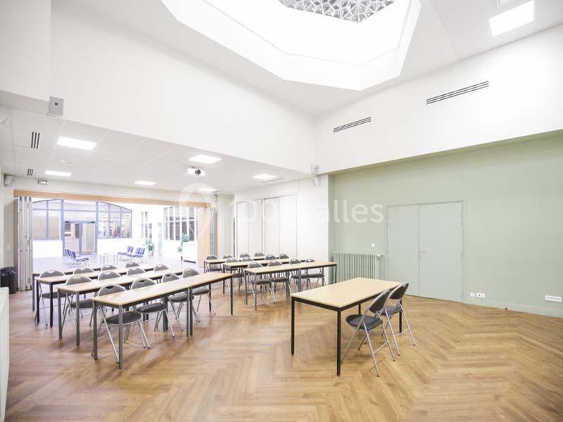 Salle lumineuse avec parquet, tables et chaises alignées, grandes fenêtres et plafond avec puits de lumière.