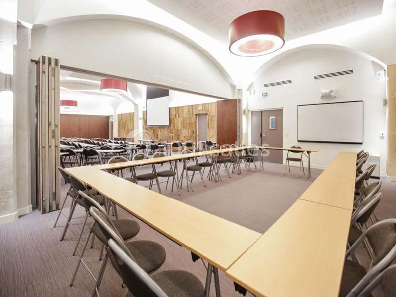 Salle de réunion lumineuse avec tables disposées en U, chaises pliantes, écran de projection et tableau blanc.