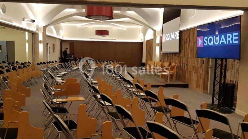 Salle de conférence avec des rangées de chaises vides, un écran affichant ’Square’ et une estrade au fond.