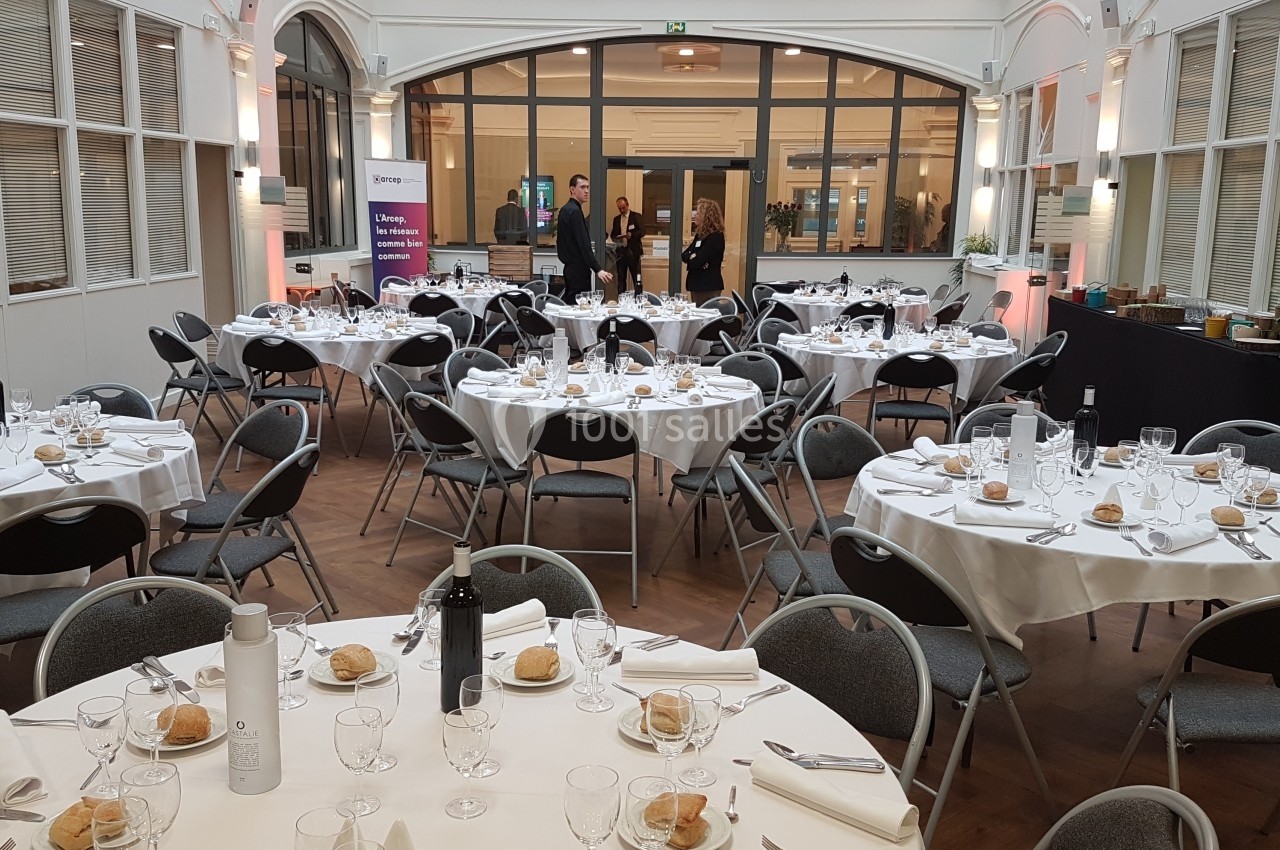Salle de réception lumineuse avec tables rondes dressées pour un repas, incluant verres, assiettes et pain.