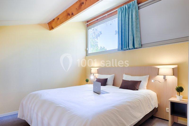 Chambre lumineuse avec un lit double, deux lampes de chevet, des coussins bruns et une fenêtre avec rideaux bleus.