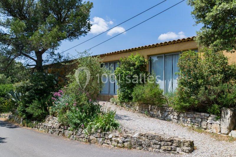 Maison en pierre avec grandes baies vitrées, entourée de végétation et située le long d'une petite route.
