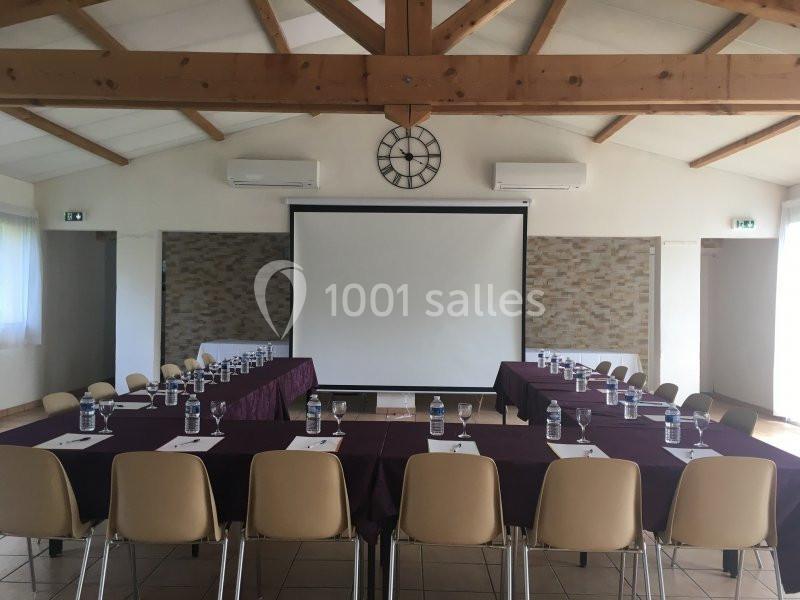 Salle de réunion lumineuse avec tables disposées en U, chaises beige, écran de projection et horloge murale.