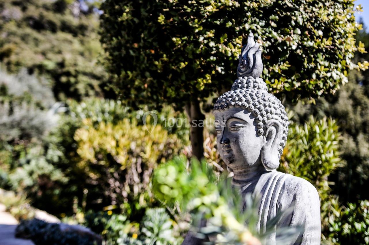 Statue de Bouddha en pierre dans un jardin verdoyant avec des arbres et des plantes en arrière-plan.