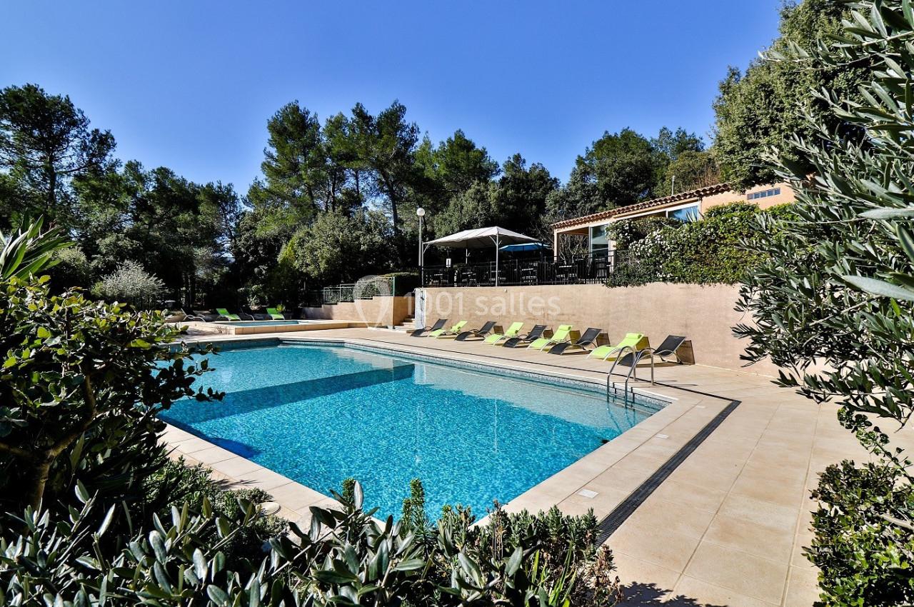 Piscine extérieure entourée de chaises longues, végétation et maison en arrière-plan sous un ciel dégagé.