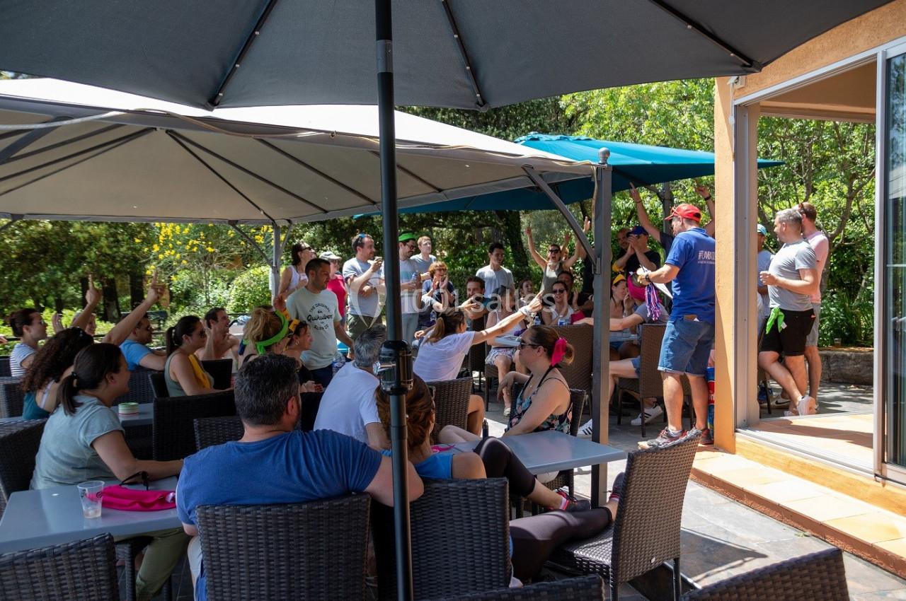 Groupe de personnes assises et debout sous des parasols, participant à une activité en extérieur près d'une terrasse.