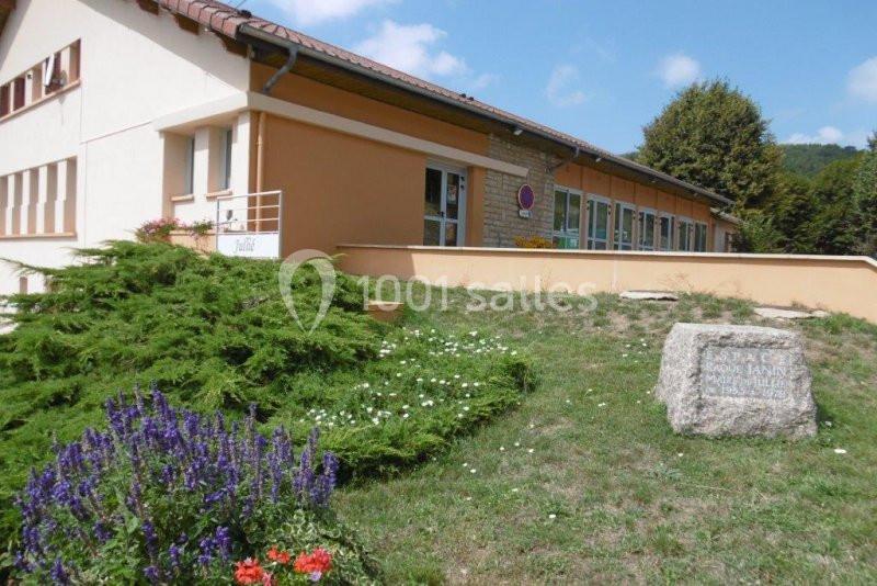Bâtiment beige avec toit en pente, entouré de verdure, parterre fleuri et pierre gravée au premier plan.