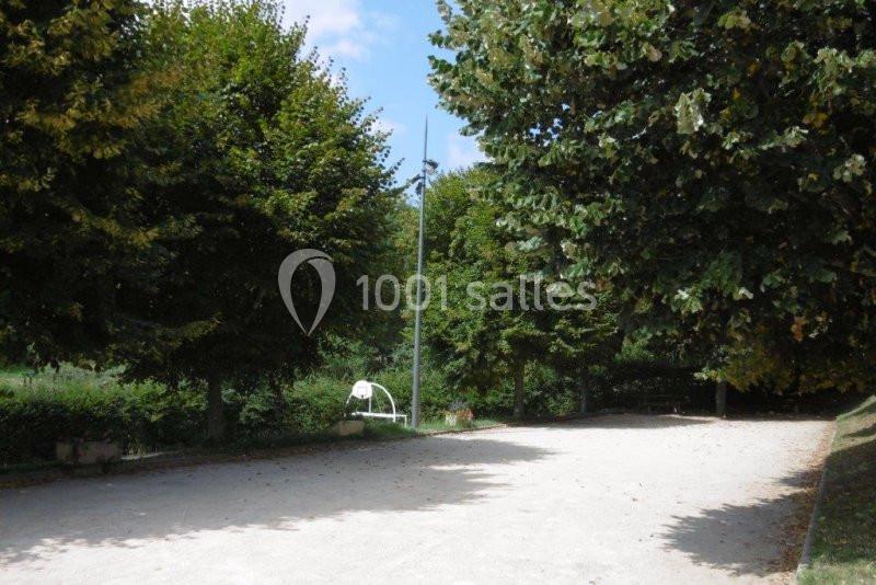 Allée bordée d'arbres avec un lampadaire central et un banc blanc sur un sol en gravier, par temps ensoleillé.